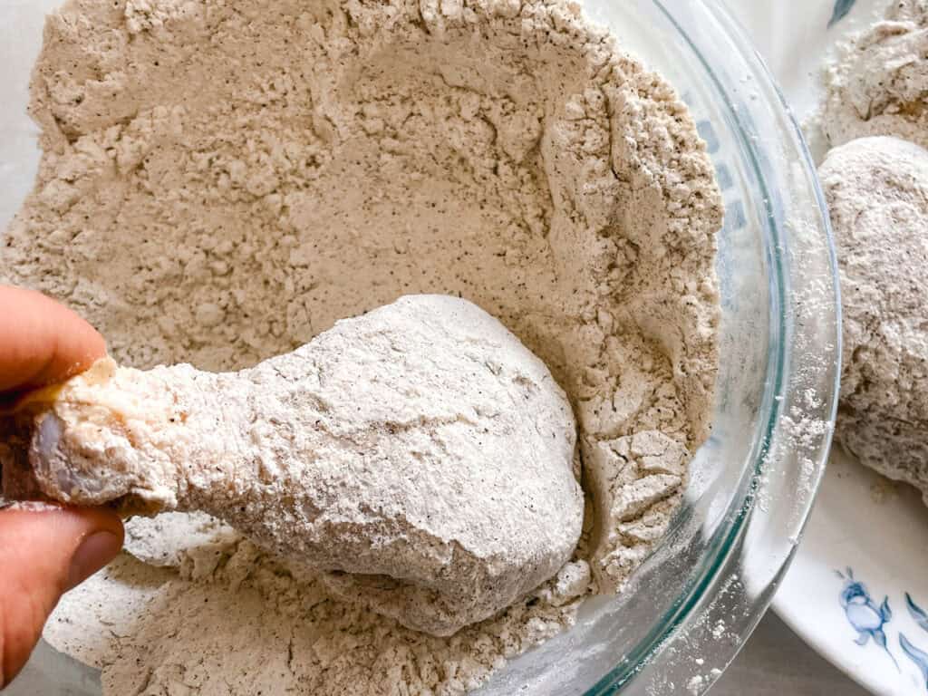 Chicken drumstick being dredged in a flour mixture before frying.