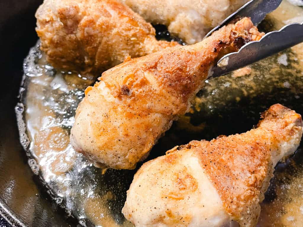 Chicken drumsticks frying in a cast iron skillet.