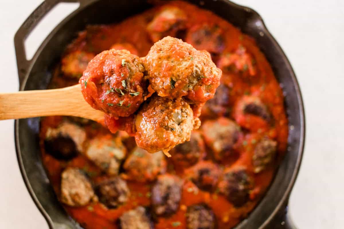 Three meatballs on a wooden spoon being held above a skillet full of meatballs and sauce.