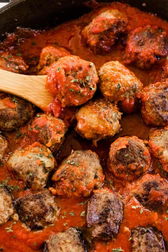 Stovetop meatballs simmering in marinara sauce in a cast iron skillet while its being stirred with a wooden spoon.
