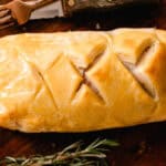 Venison wellington with a flakey breading and beautiful cut marks on top sitting on a wooden cutting board.