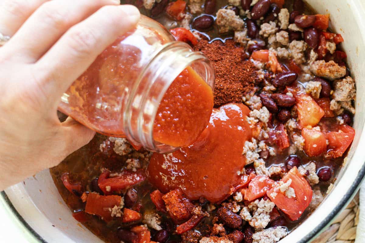 Pouring tomato sauce into a dutch oven over a ground pork and beans mixture.
