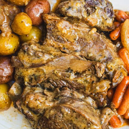Pot roast with gravy, new potatoes, and baby carrots on white serving platter with a checkered towel under it.