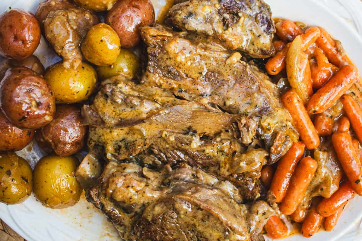 Pot roast with gravy, new potatoes, and baby carrots on white serving platter with a checkered towel under it.