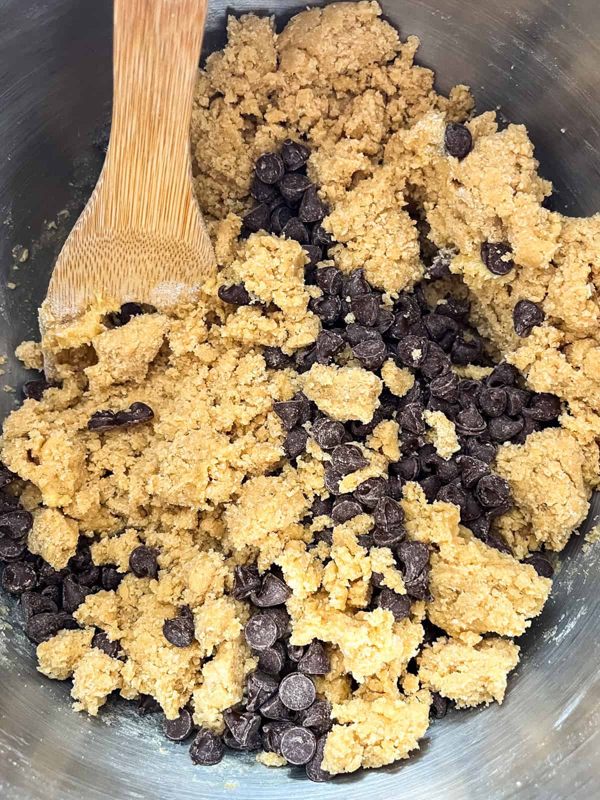 Chocolate chips being mixed into cookie dough with a wooden spoon.