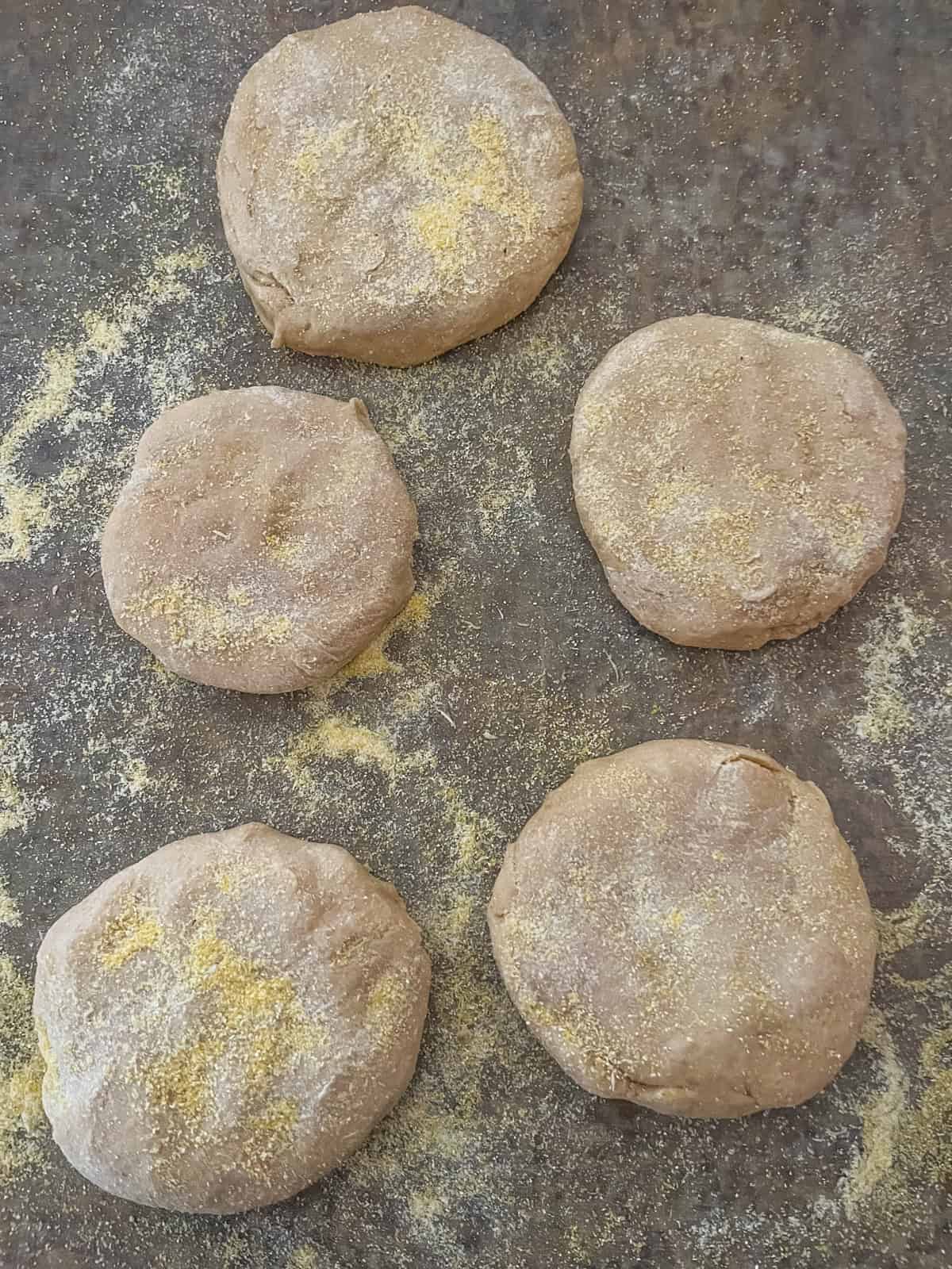 Uncooked English muffins cut out with cornmeal sprinkled over the dough and counter.