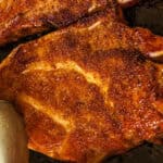 Flipping the pork steak with a pair of tongs on a cast iron skillet.