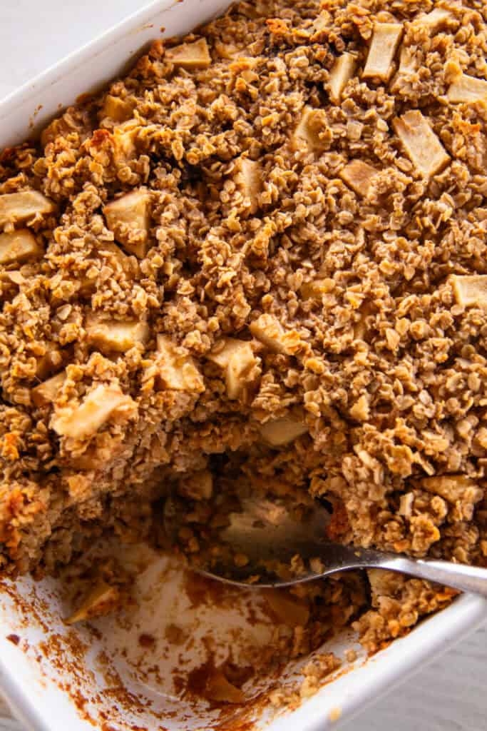 A metal spoon scooping out Apple Cinnamon Oatmeal Bake from a white baking dish.