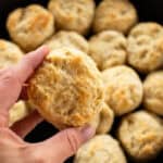 A single Cast Iron Biscuit being held above a skillet filled with biscuits.
