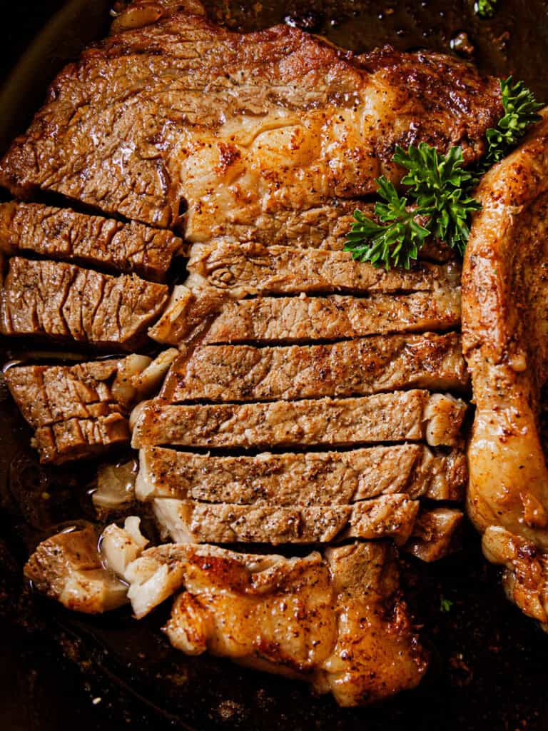 Ribeye steak sitting in a cast iron skillet, freshly seared and sliced, with a fresh herb garnish.