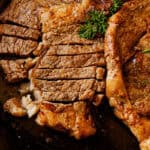 Two seared ribeye steaks sitting in a cast iron skillet, one of them sliced, with a fresh herb garnish.
