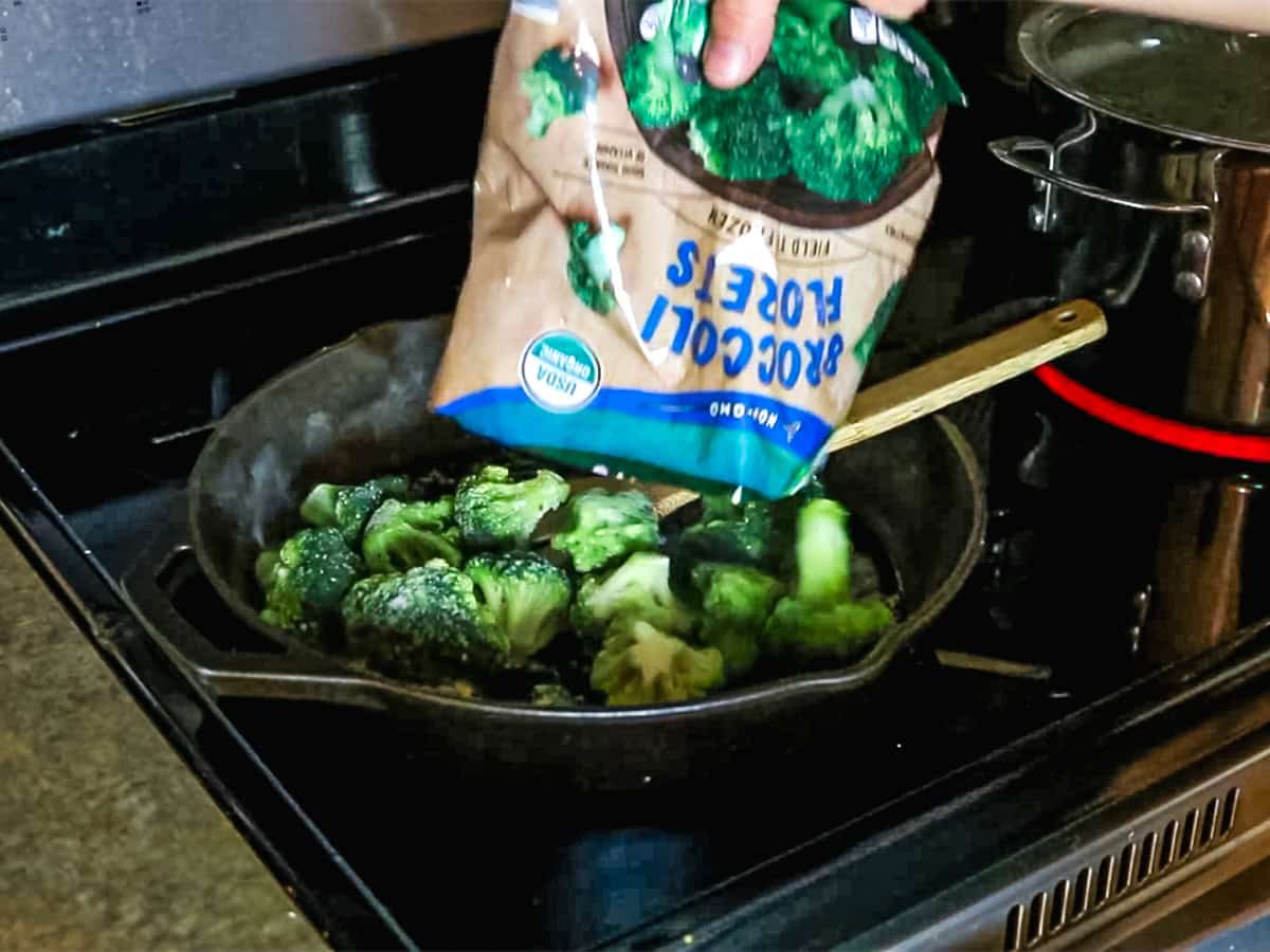 Pouring a bag of frozen broccoli into a cast iron skillet on the stove.