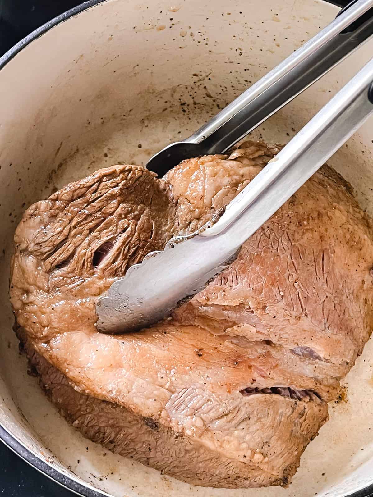 Searing a brisket in a dutch oven, using metal tongs to hold the meat.