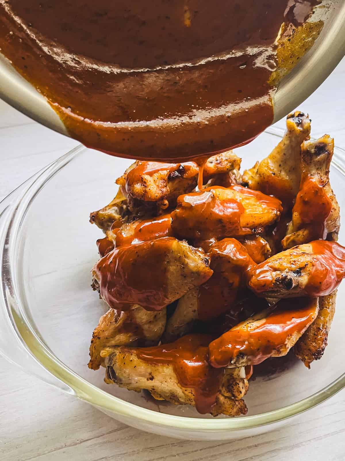Pouring wing sauce on baked wings in a glass bowl.
