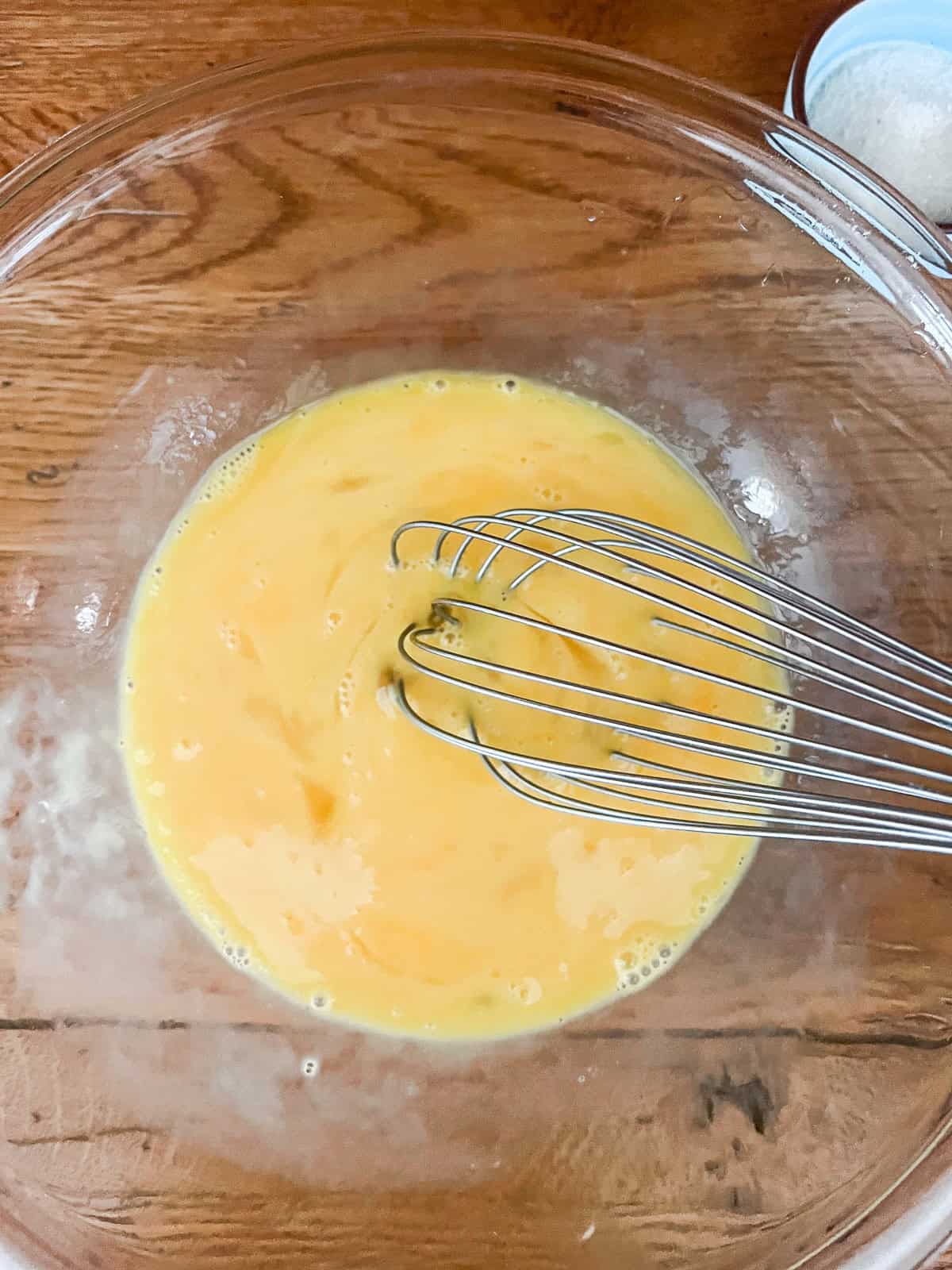 Fully mixed eggs in a glass bowl, with a metal whisk.