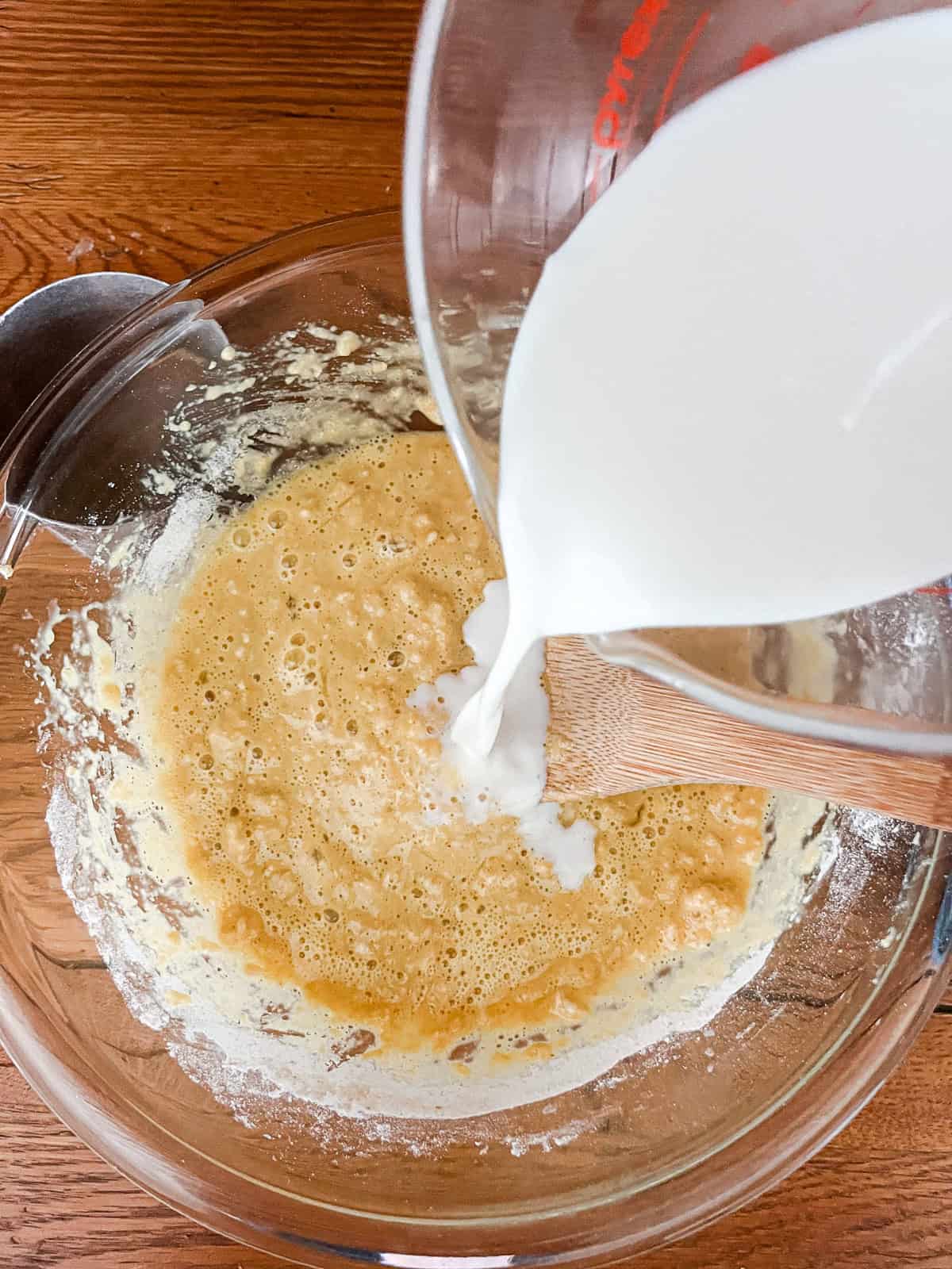 Pouring milk into a flour and egg mixture in a glass bowl.