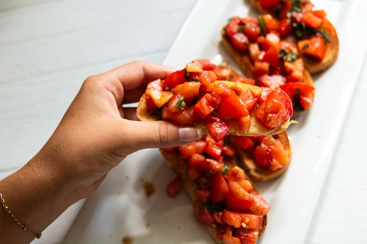 Holding a piece of bruschetta up over a plate of more bruschetta.