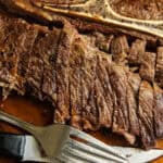 A sliced pan-seared chuck steak on a wooden board with a fork and knife.