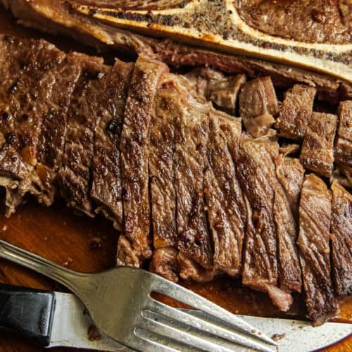 A sliced pan-seared chuck steak on a wooden board with a fork and knife.