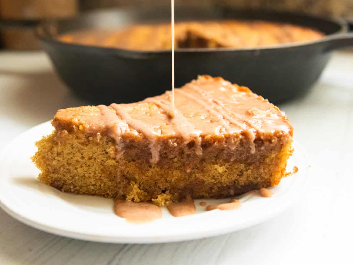 Drizzling glaze on a slice of apple cider cake on a white plate. 