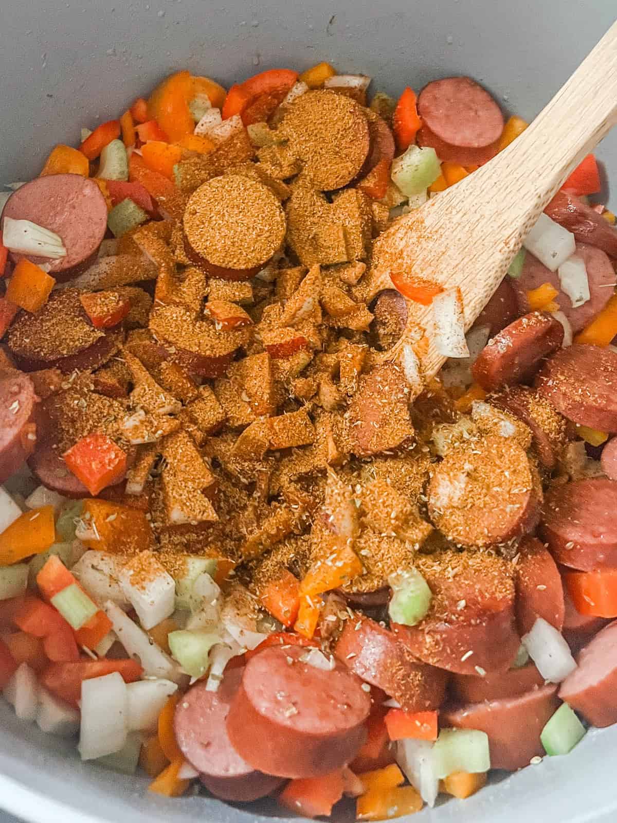 Sauteed veggies, sausage, and seasoning in a Dutch oven, being stirred with a wooden spoon.