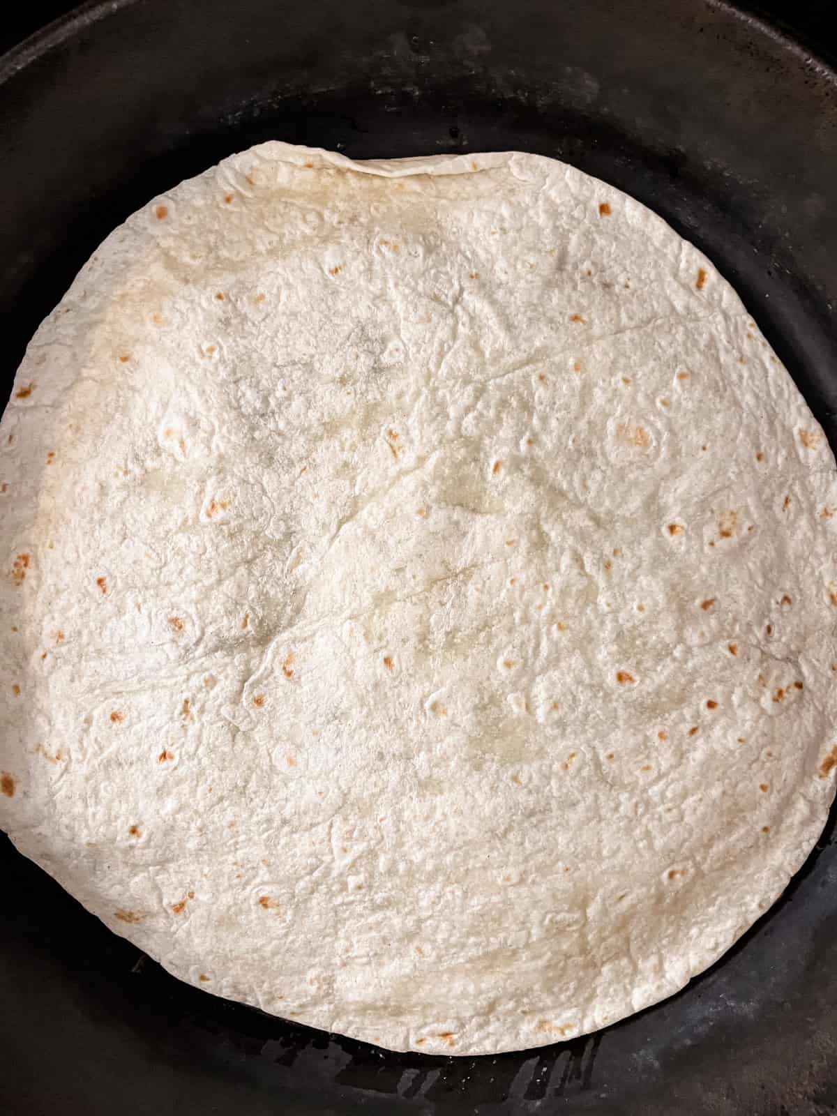 A tortilla on top of a quesadilla, cooking in a cast iron skillet.