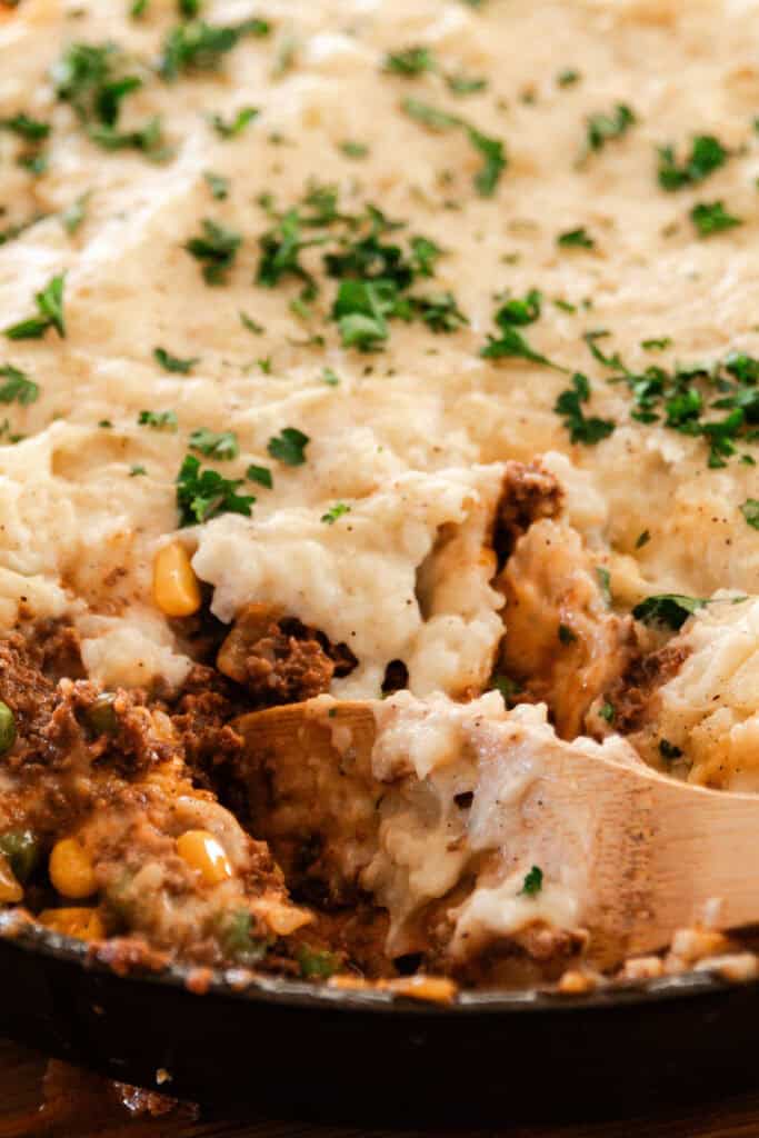 Scooping venison shepherd's pie out of a cast iron skillet with a wooden spoon.
