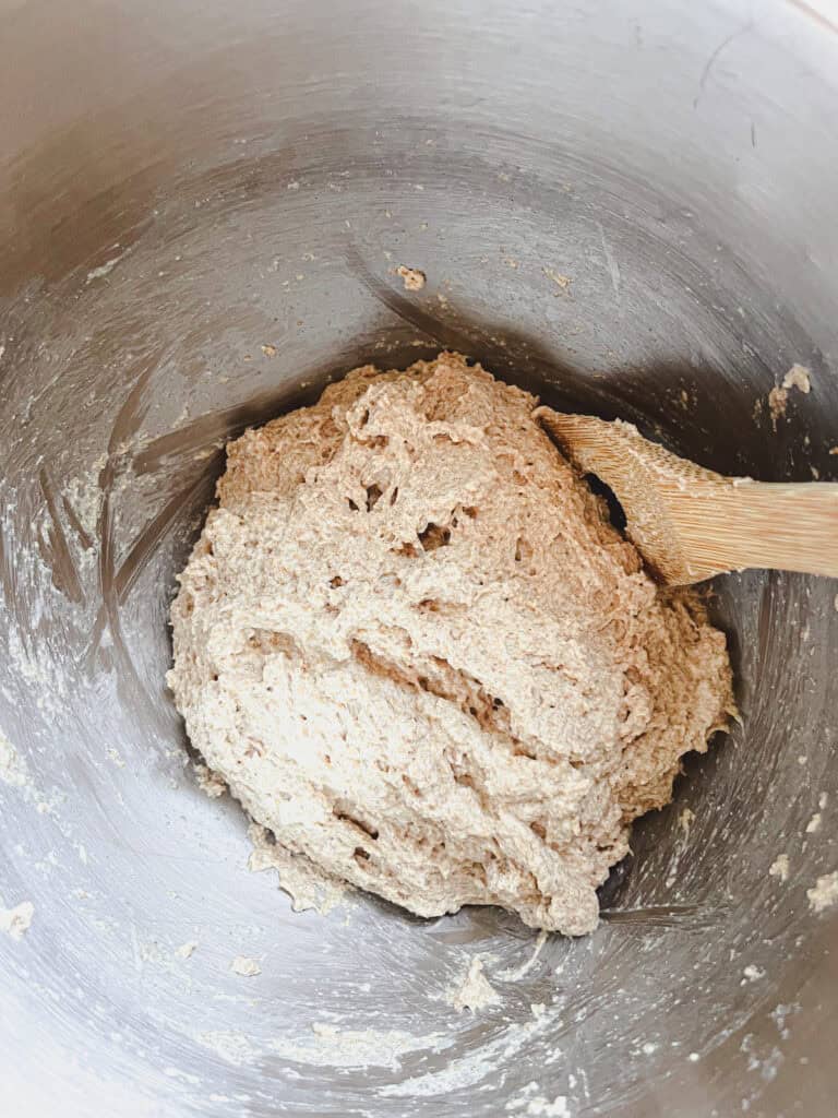 Stirring up baguette dough with a wooden spoon in a metal mixing bowl.