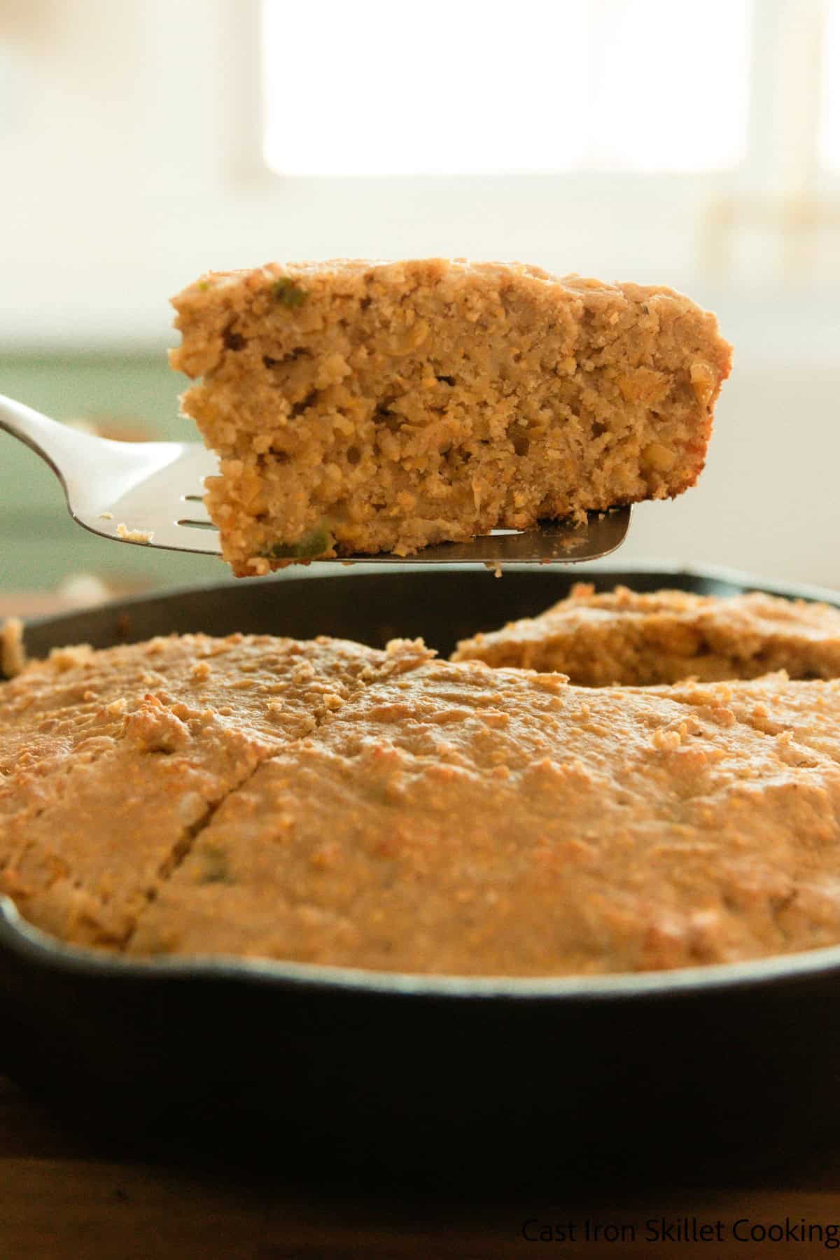 Lifting a slice of cornbread with freshly milled flour out of a cast iron skillet with a spatula.