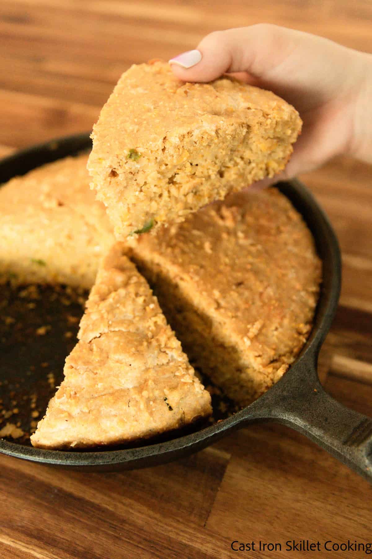 A hand lifting a spice of freshly milled cornbread out of a cast iron skillet.