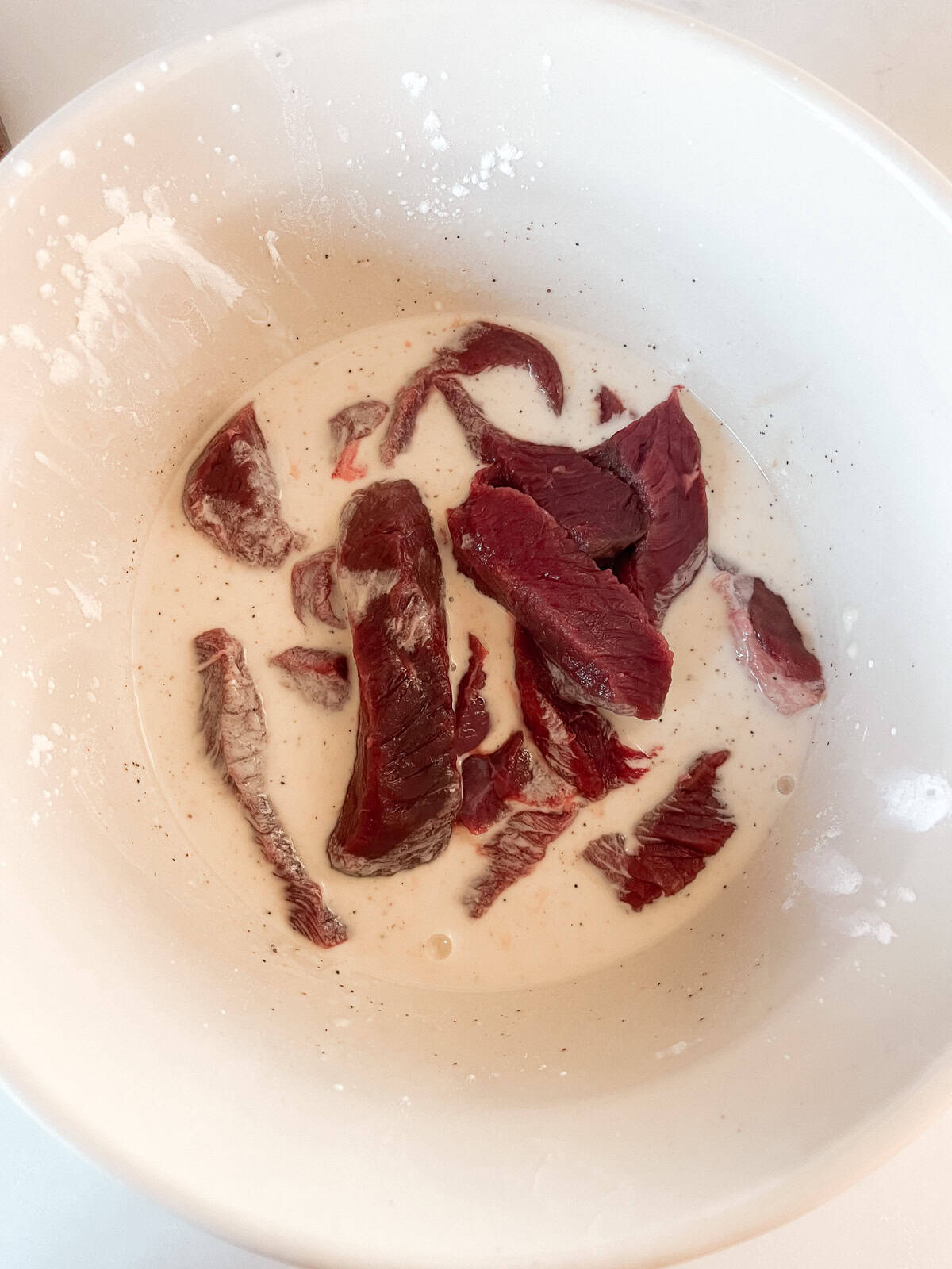 Venison steak strips in a bowl of flour and egg mixture.