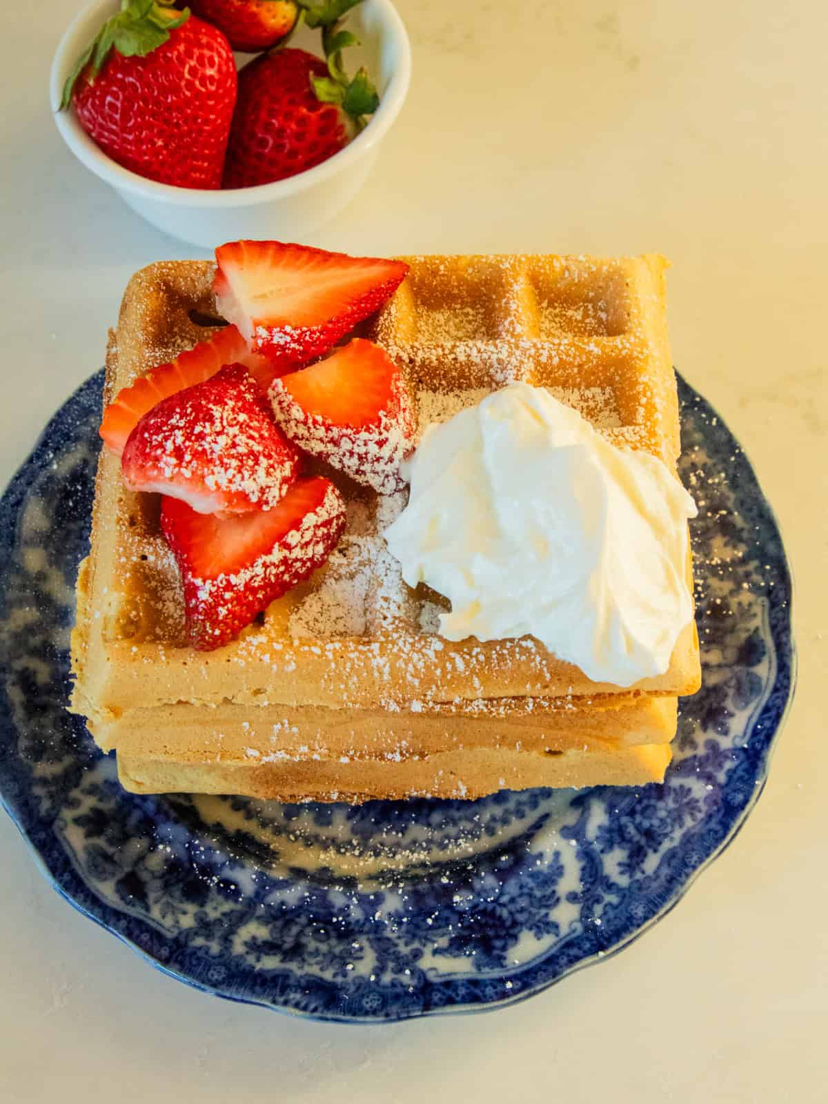 3 freshly milled waffles on a blue decorative plate with fresh strawberries and homemade whipped cream.