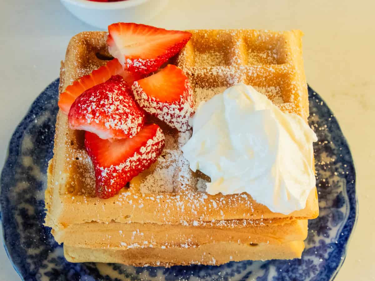 Tall stack of freshly milled waffles with fresh strawberries and whipped cream sprinked with powdered sugar.
