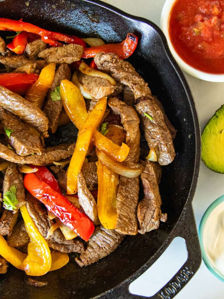 A cast iron skillet with venison fajitas in it with sour cream, avocado, and salsa on the side.
