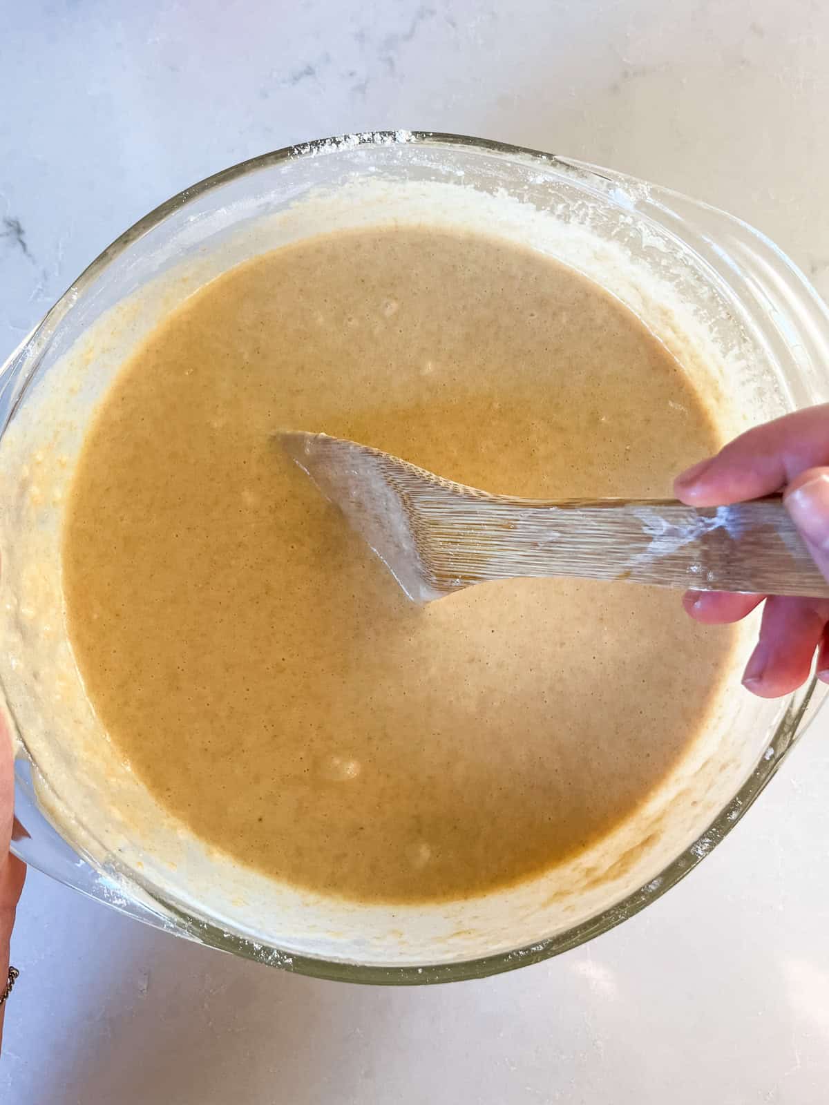 Stirring fresh milled waffle batter in a glass bowl with a wooden spoon.