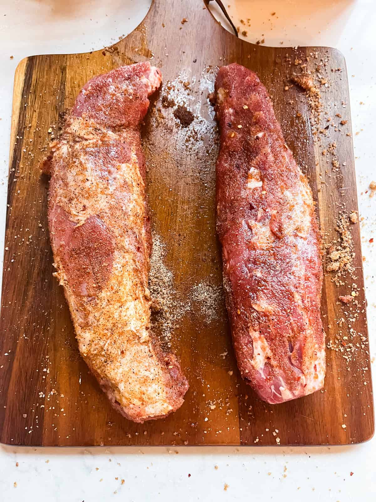 Pork tenderloin on a wooden cutting board, sprinkled with rub.