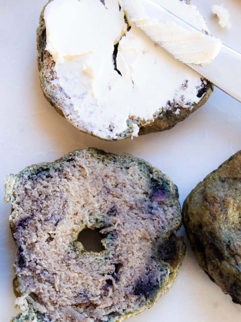 A freshly milled blueberry bagel smeared with cream cheese with more bagels next to it.