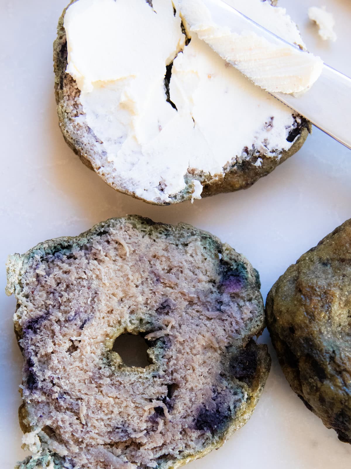 A freshly milled blueberry bagel smeared with cream cheese with more bagels next to it.
