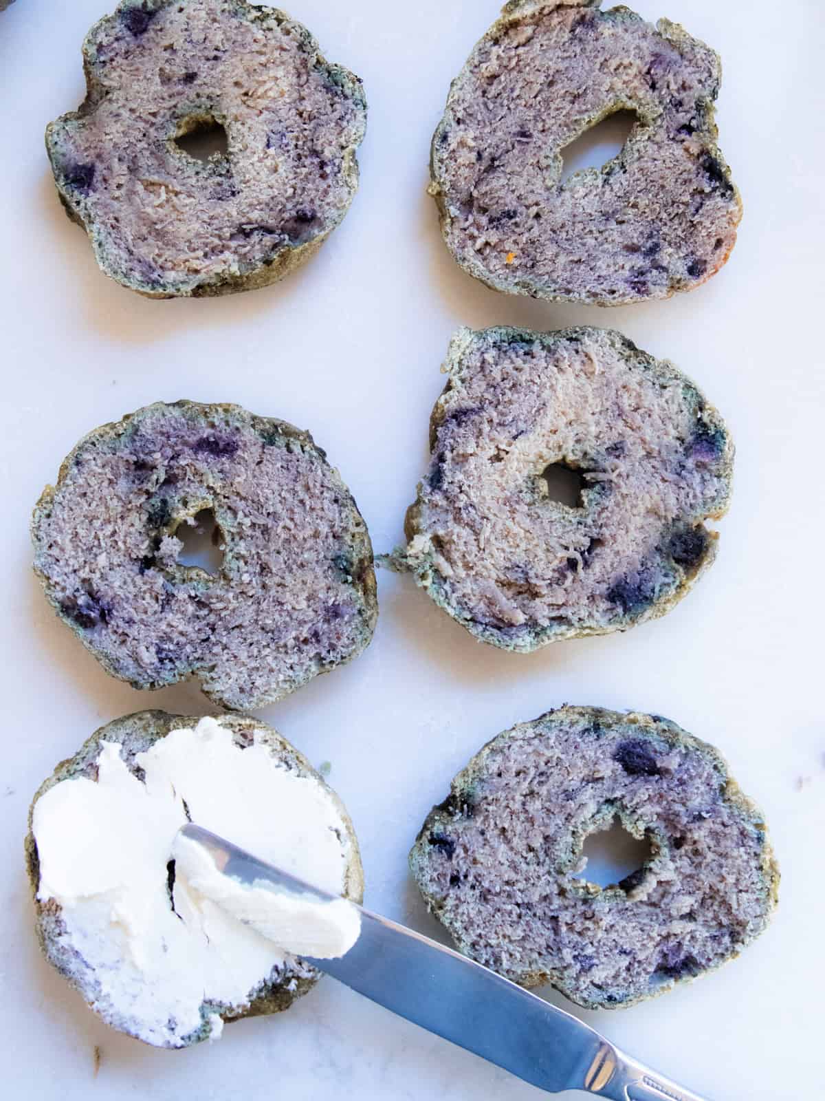 Blueberry bagels lined up on a kitchen counter, one with cream cheese spread on it.
