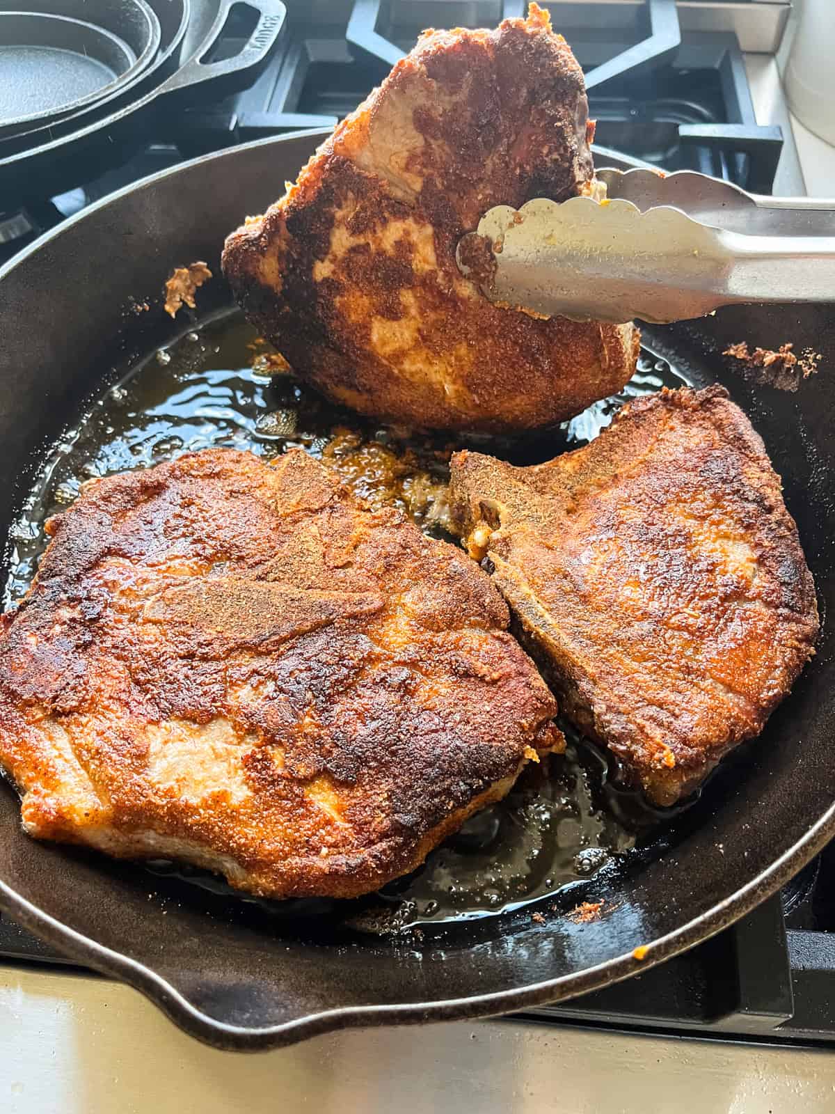 Frying pork chops in a cast iron skillet and flipping one with tongs.