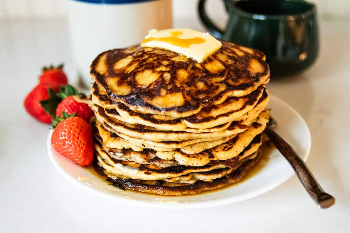 Whole wheat pancakes with freshly milled flour on a serving plate with a gold fork, large dollop of butter and syrup dripping off the sides.