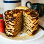 Whole wheat pancakes with freshly milled flour stacked in a tall pile on a plate with butter on top and maple syrup falling down the sides and some fresh strawberries next to it.