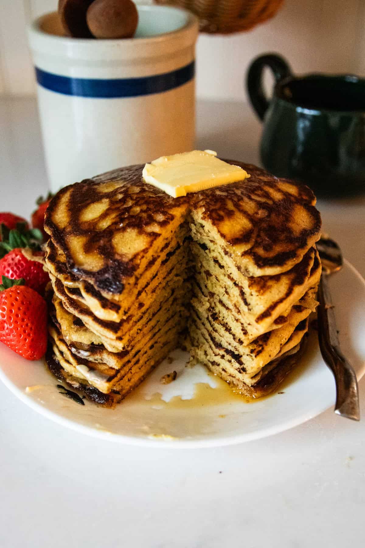 Tall stack of freshly milled flour pancakes on a plate with butter on top and maple syrup falling down the sides and some fresh strawberries next to it. There is also a big slice missing out of the middle.