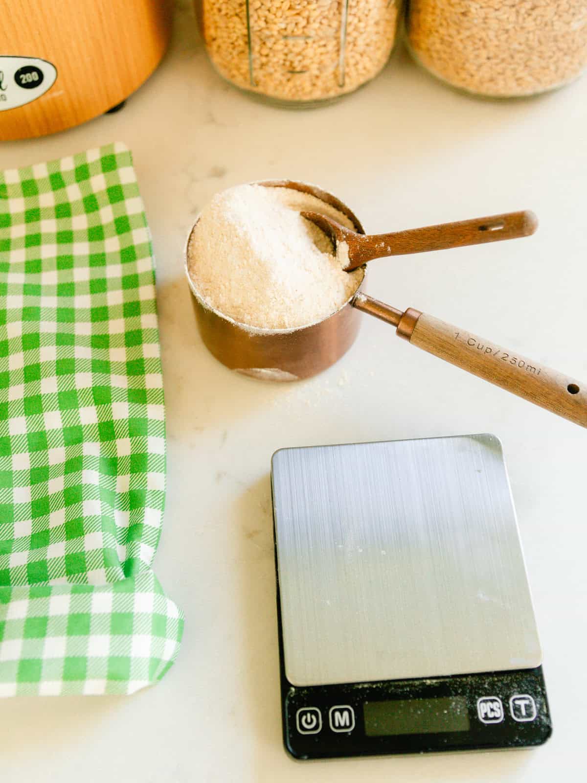 A measuring cup of freshly milled flour with a kitchen towel and a digital scale.