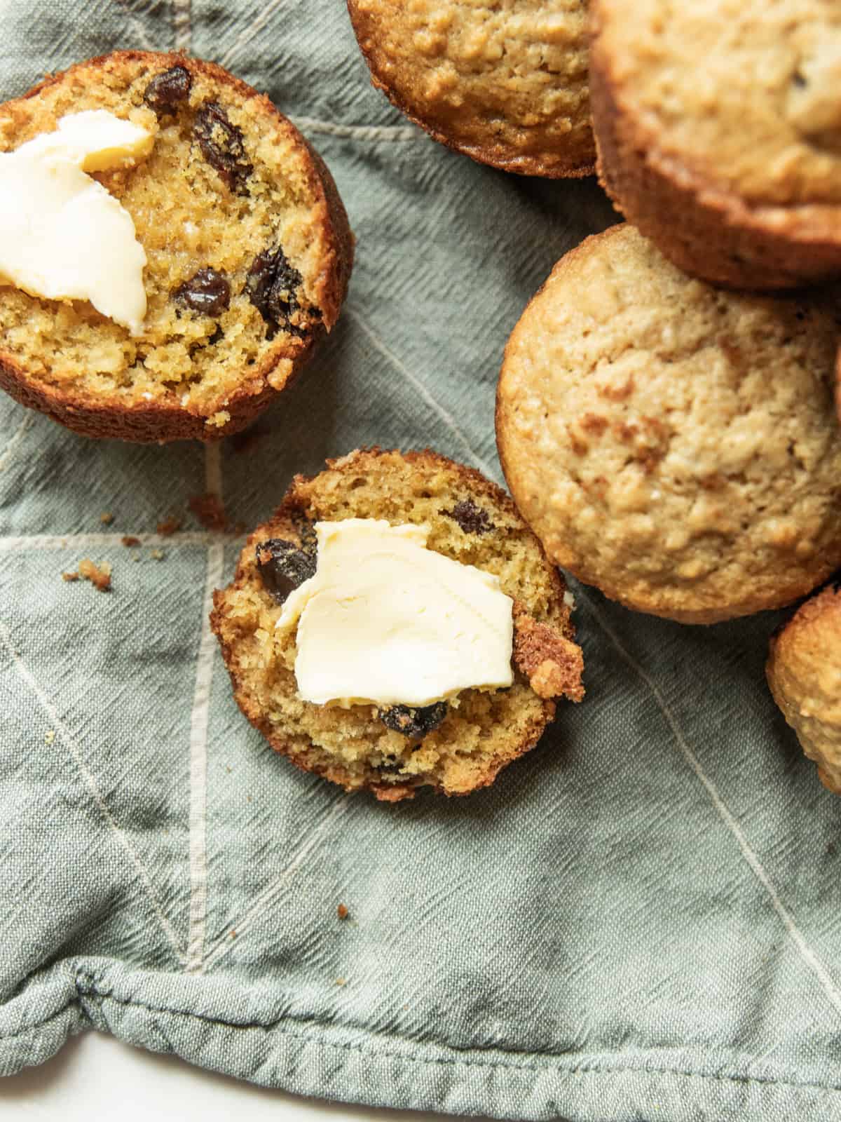 An oatmeal raisin muffin cut in half with butter on it with more muffins on the side.