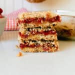 A stack of strawberry crumble bars with a red checkered napkin and a bowl of strawberries in the background.