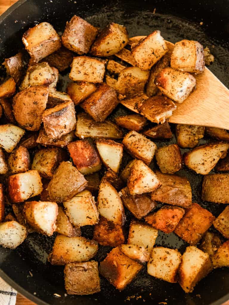 Scooping cast iron skillet potatoes out of a skillet with a wooden spoon.