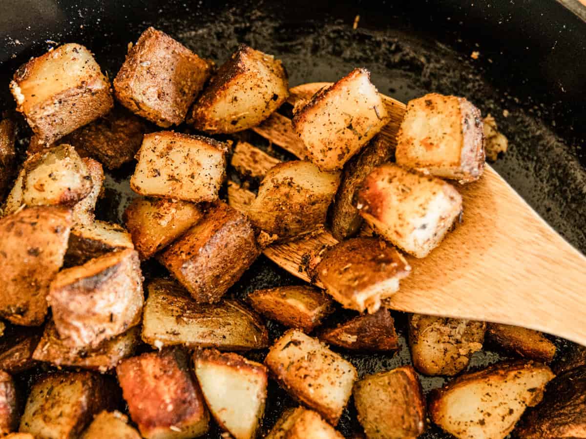 Scooping cast iron skillet potatoes out of a skillet with a wooden spoon.