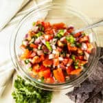A bowl of pico de gallo with a scoop of tortilla chips and a pinch of cilantro on the side.