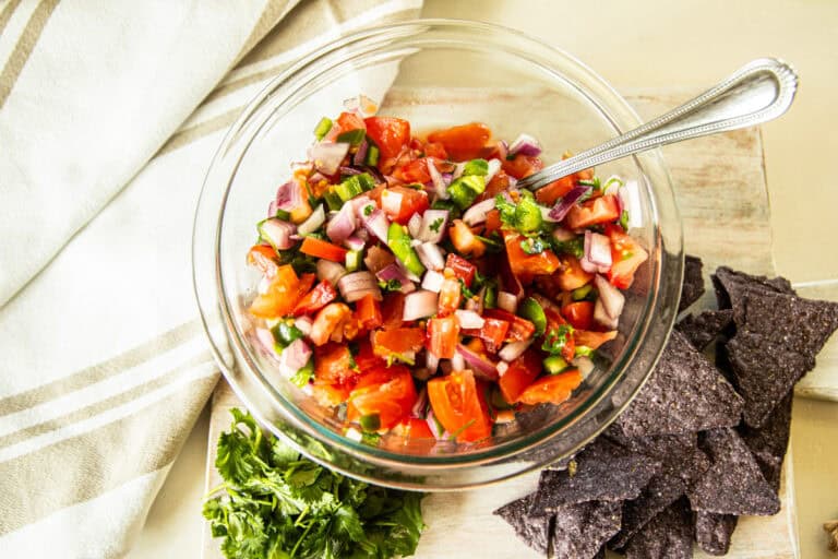 A bowl of pico de gallo with a scoop of tortilla chips and a pinch of cilantro on the side.